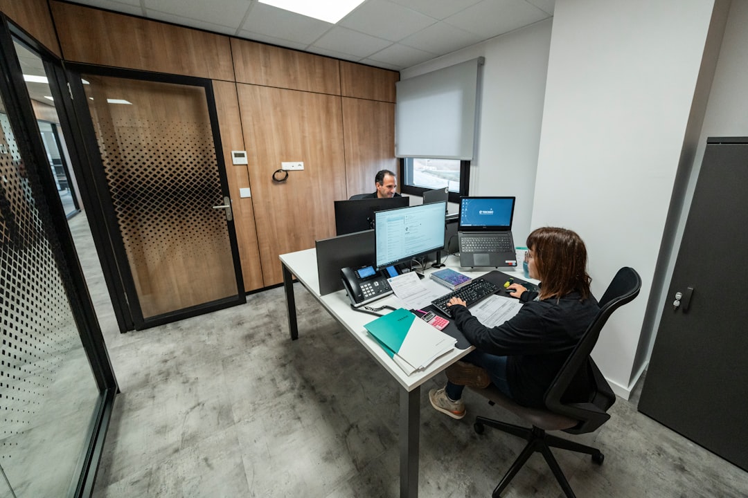 Modern smart office workspace with professional employee working at computer desk with dual monitors, organized documents, phone system, and contemporary office interior with glass partitions